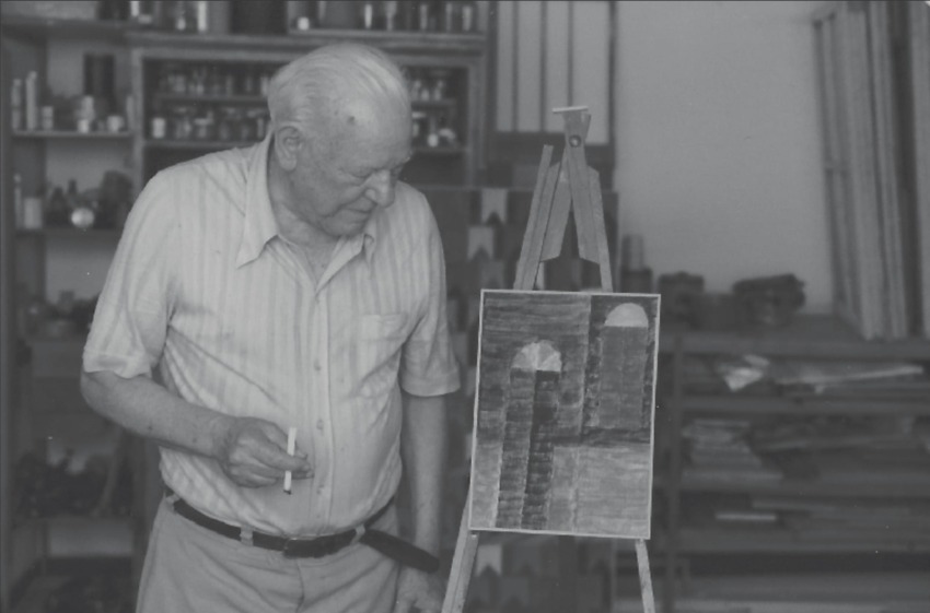 Elderly man painting in a cluttered studio, focusing on a small canvas on an easel with geometric shapes.