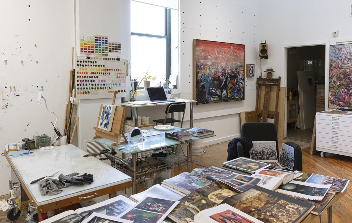 Artist's studio with paintings, color swatches, tables, and art supplies, featuring natural light from a large window.