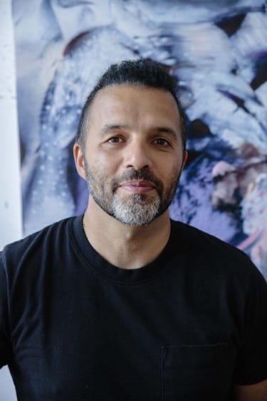 Man in a black t-shirt standing in front of an abstract painting, looking at the camera.