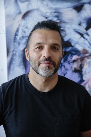 Man in a black t-shirt standing in front of an abstract painting, looking at the camera.