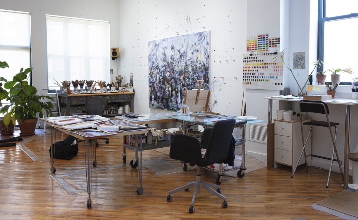 Artist's studio with wooden floor, desks with art supplies, large painting on wall, and potted plants by the windows.