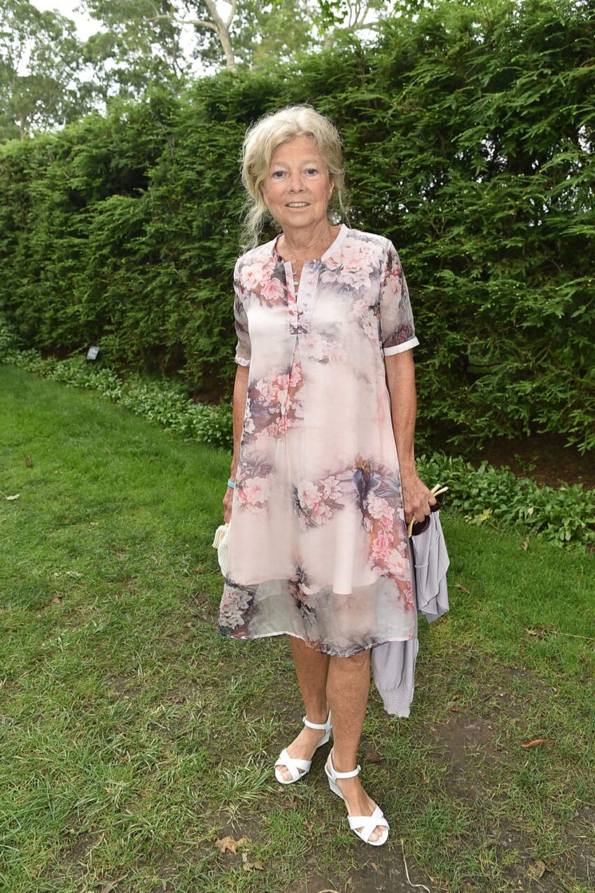 An individual in a floral dress stands on grass with a hedge in the background.