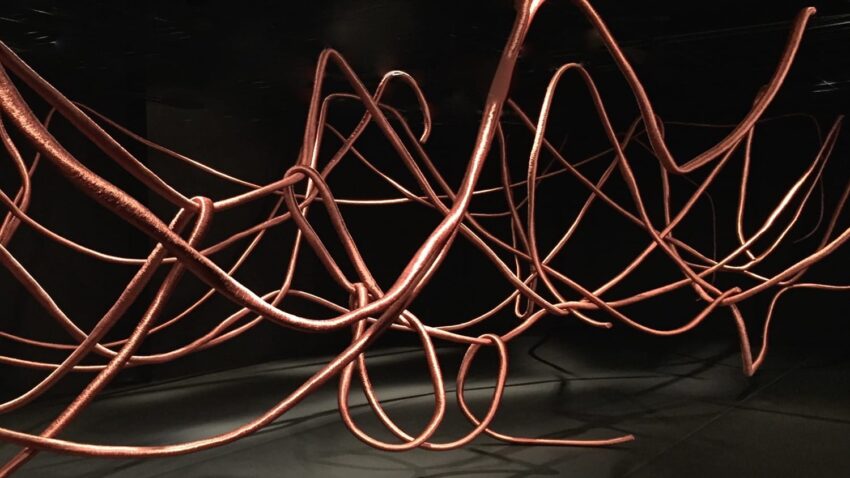 Abstract installation with intertwined, copper-colored wires suspended in a dark room creating a chaotic, artistic effect.
