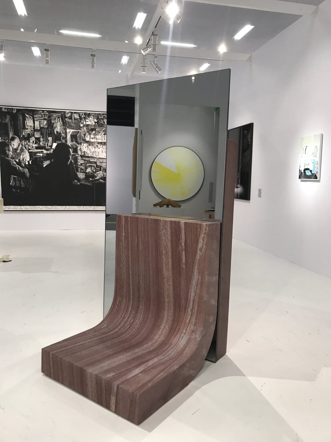 Modern art sculpture in a gallery with a curved stone base and reflective glass, surrounded by black and white photos.