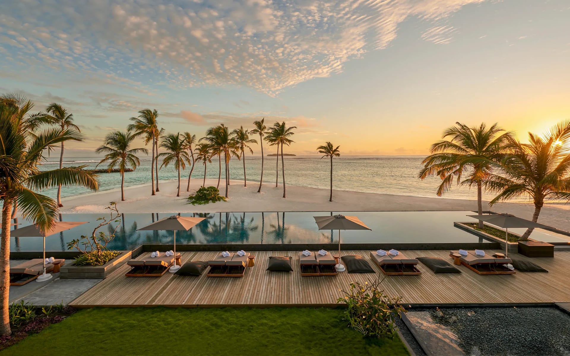 Luxury resort infinity pool overlooking the ocean at sunset, surrounded by palm trees and sun loungers on wooden deck.