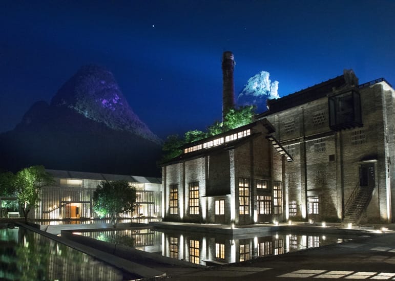 Industrial building with brick chimney and lit windows at night, mountains in background, reflected in calm water surface.