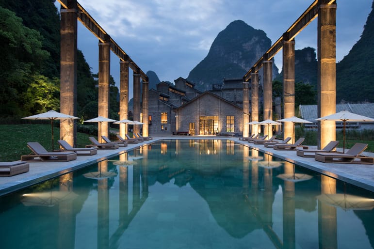 Luxurious resort pool with loungers and umbrellas, surrounded by tall pillars and mountainous landscape at dusk.