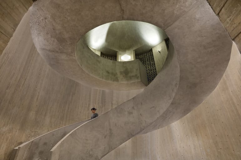 Person ascending a spiral staircase inside a modern, concrete building with dramatic architectural features.