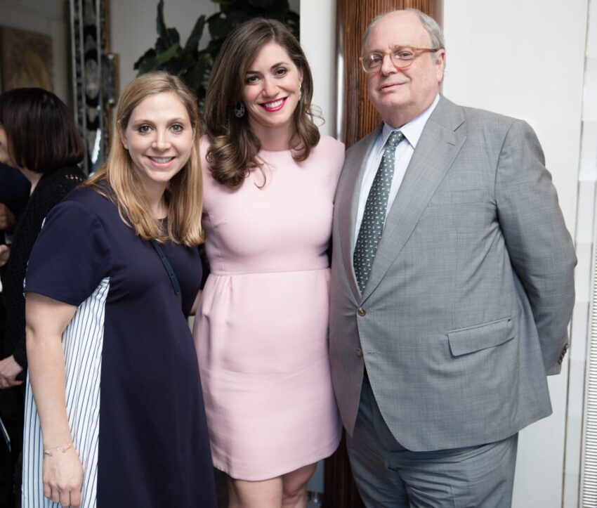 Three people smiling together, two women in dresses and a man in a suit, standing in a social setting indoors.