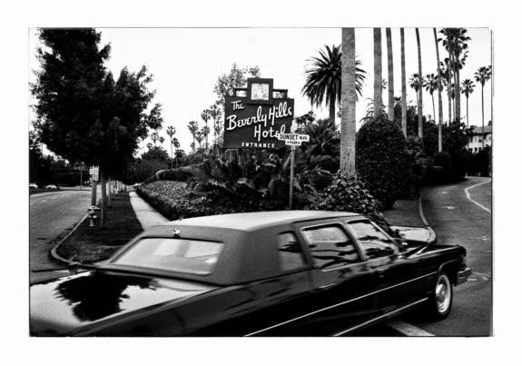 Black and white photo of a vintage car passing the Beverly Hills Hotel sign surrounded by tall palm trees.