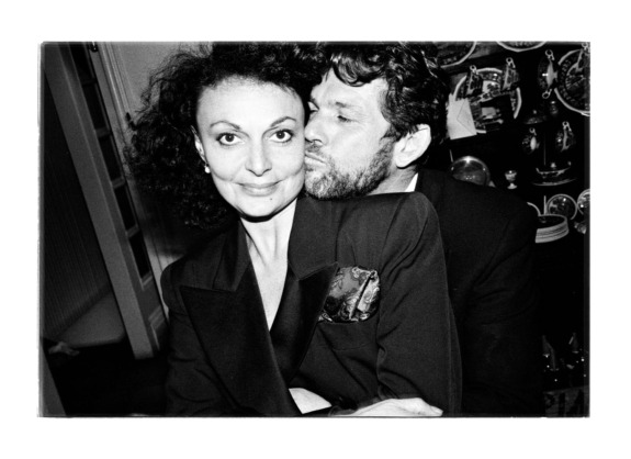 Man embraces woman affectionately while she smiles, both dressed in formal attire indoors, with decorative plates in the background.
