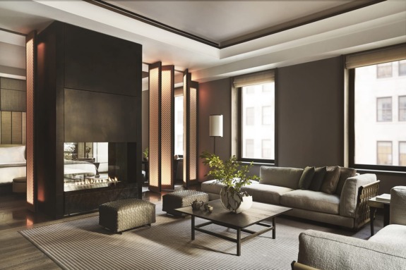 Modern living room with gray sofas, a coffee table, and a central fireplace, illuminated by natural light from large windows.