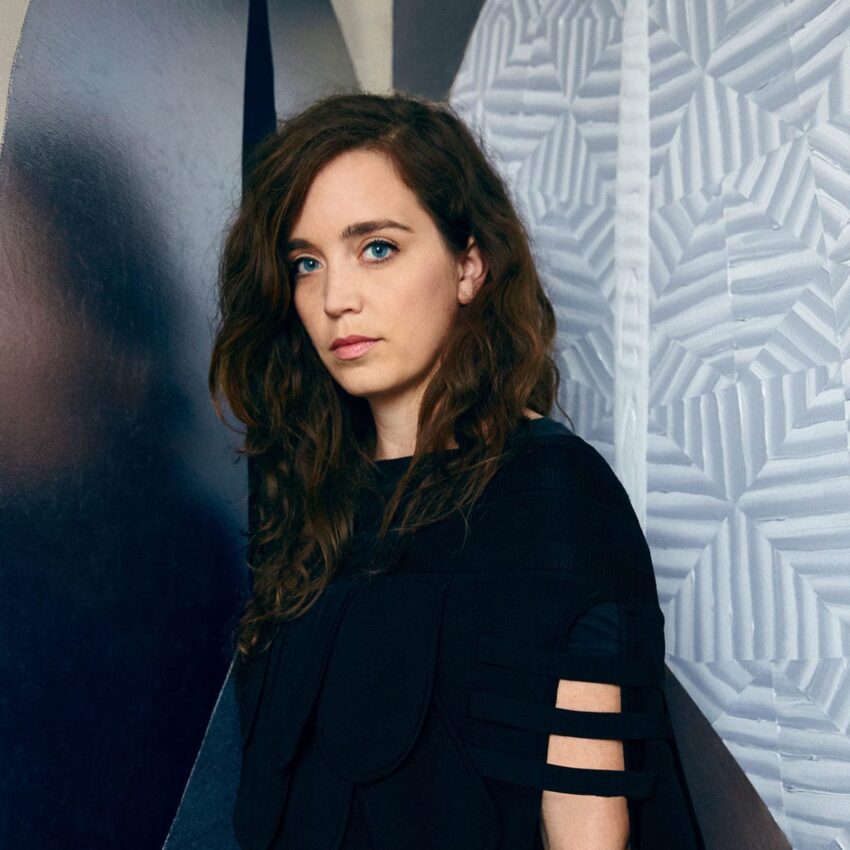 Woman with long brown hair in a black dress standing against a textured wall background.