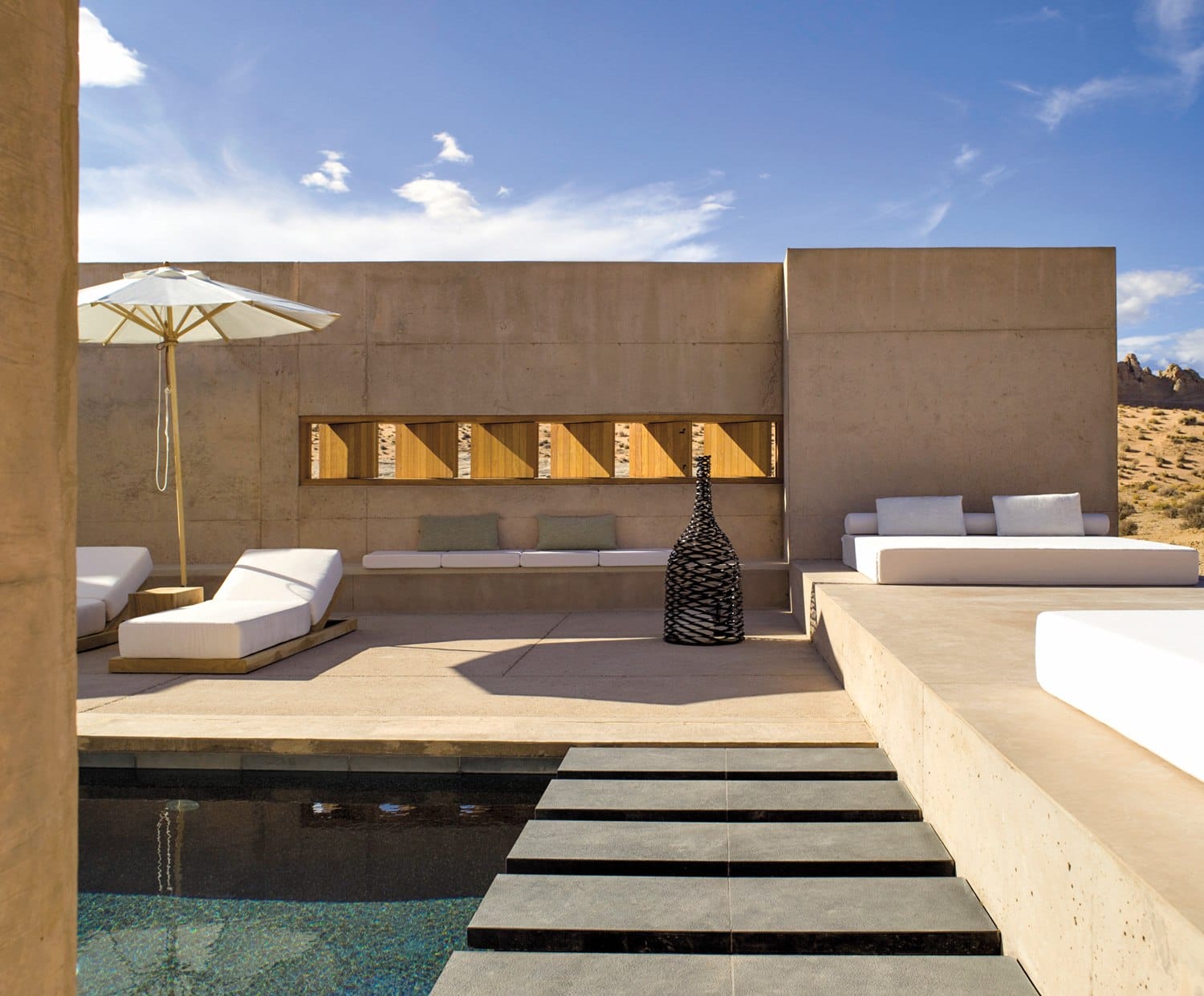 Modern outdoor patio with white loungers, pool, and geometric concrete walls under a blue sky.
