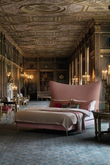 Spacious ornate room with a large pink bed, intricate ceiling, and classic decor illuminated by elegant candle-style lights.