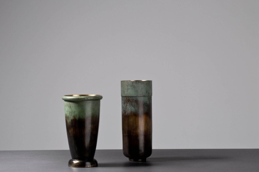 Two ceramic vases with a green and brown patina finish displayed against a gray background on a dark surface.