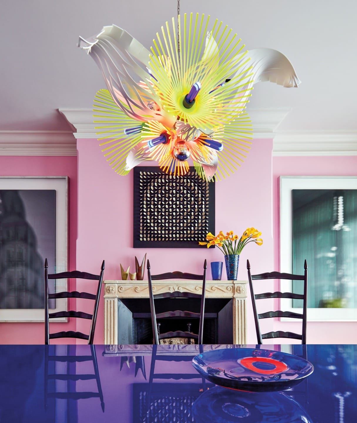 Colorful dining room with vibrant chandelier, blue table, pink walls, and artistic decor elements.
