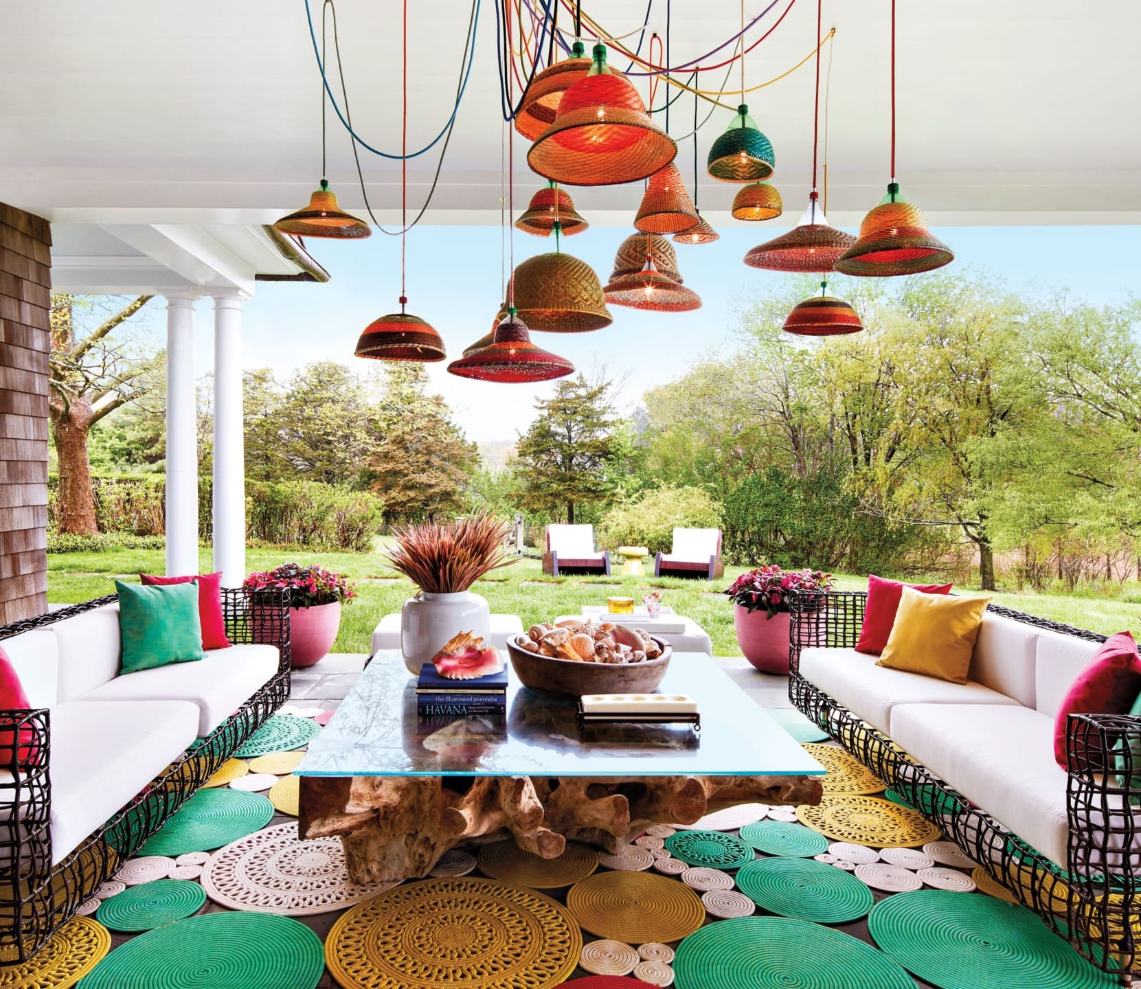 Colorful outdoor patio with vibrant hanging lamps, white sofas, and a glass table, overlooking a lush green garden.