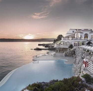 Infinity pool overlooking a calm sea at sunset with Mediterranean-style building in the background.
