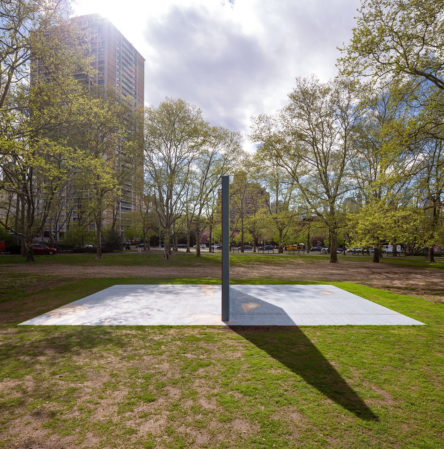 Art installation in a park with a tall structure casting a shadow on a square platform, surrounded by trees and a tall building.
