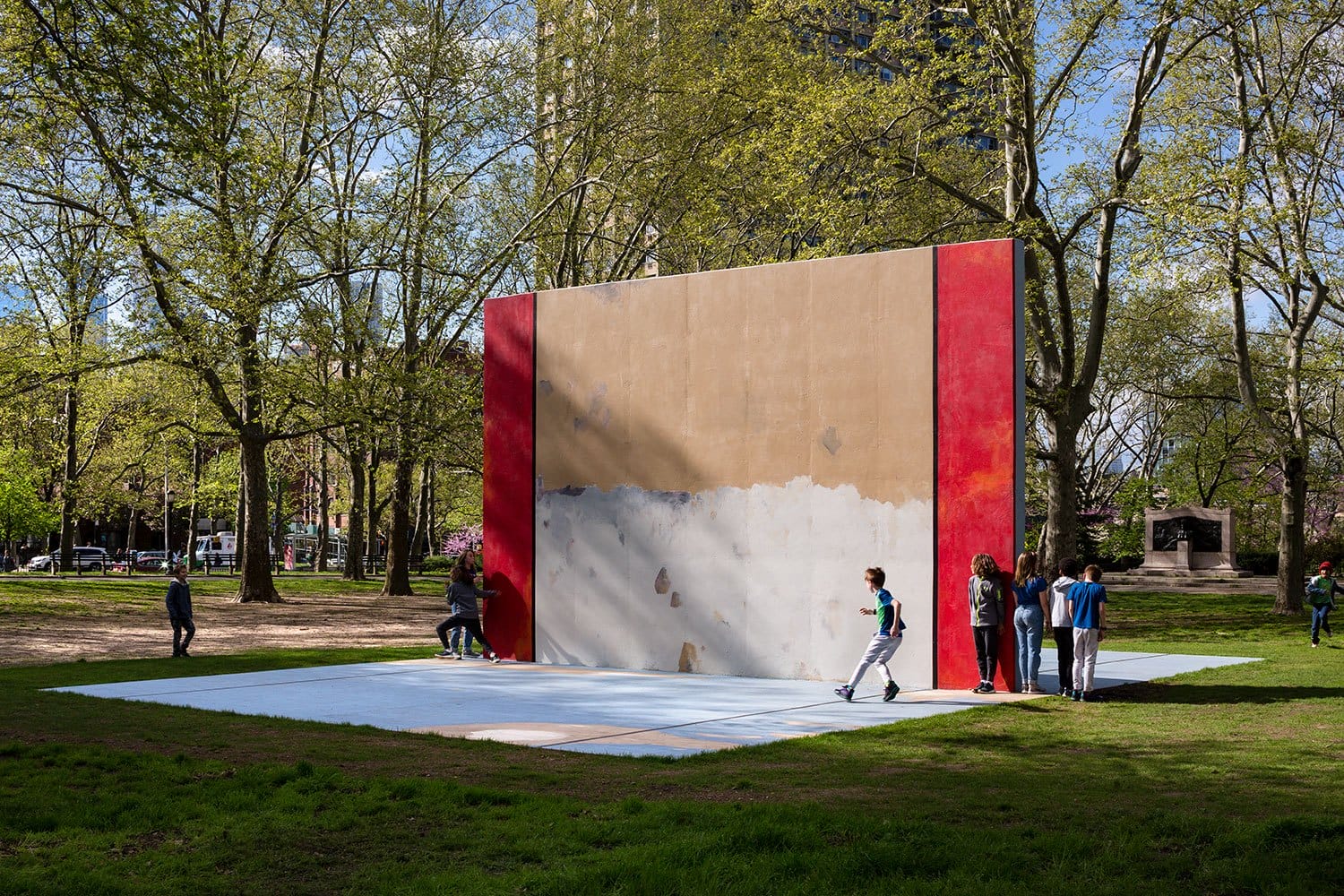 People stand near a large outdoor wall in a park, surrounded by trees, on a sunny day.
