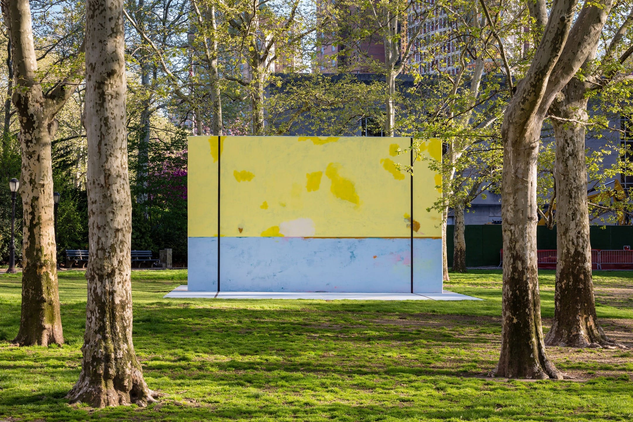 Colorful yellow and blue mural on outdoor wall among trees in a park with sunlight filtering through leaves.