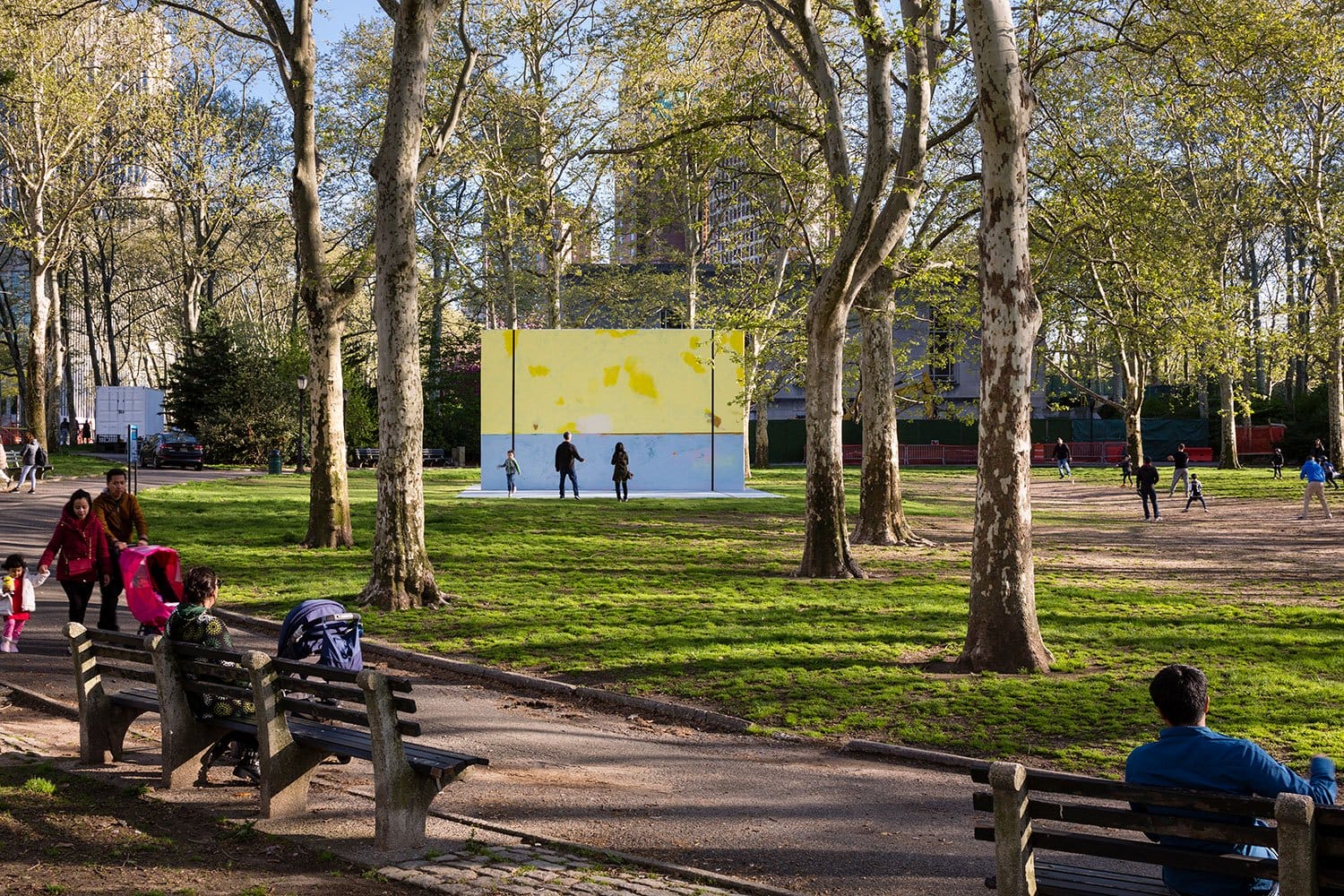 People enjoying a sunny day in a park with trees, benches, and a large painted wall in the background.