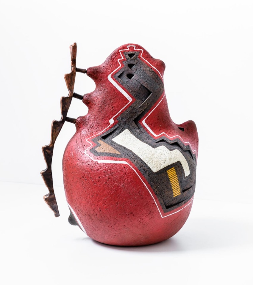 Abstract red ceramic sculpture with geometric patterns and jagged metal elements on a white background.
