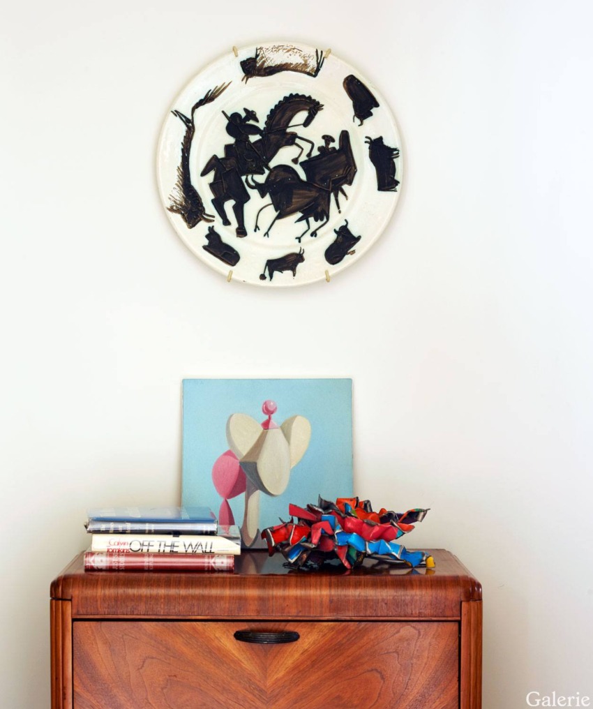 Mid-century modern wooden cabinet with art pieces, including a circular wall hanging and colorful sculpture on top.