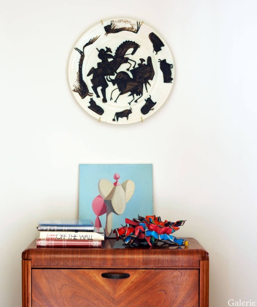 Mid-century modern wooden cabinet with art pieces, including a circular wall hanging and colorful sculpture on top.