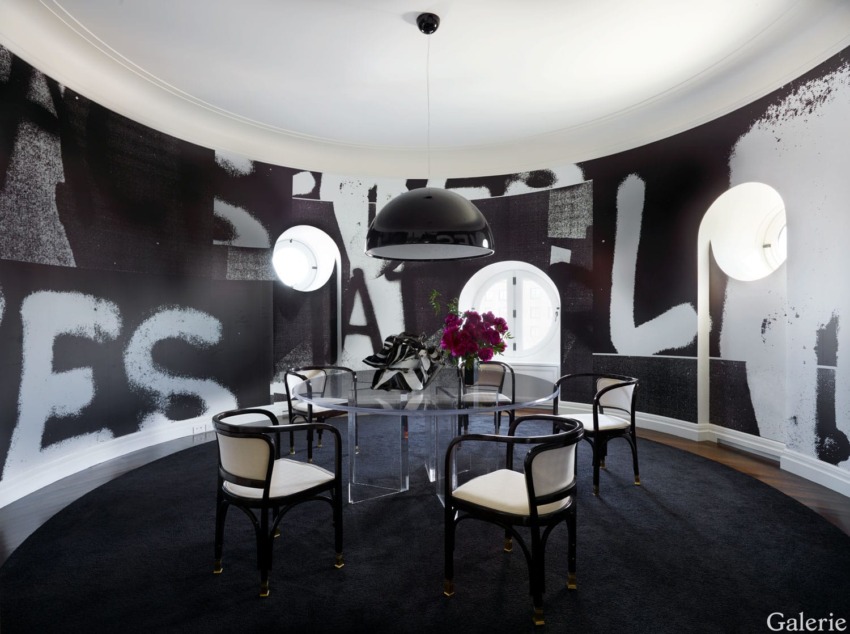 Modern dining room with circular glass table, black chairs, bold monochrome walls, ceiling light fixture, and floral centerpiece.