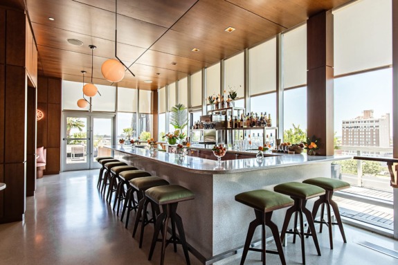Modern rooftop bar with long counter, green stools, and city view through large windows during daytime.