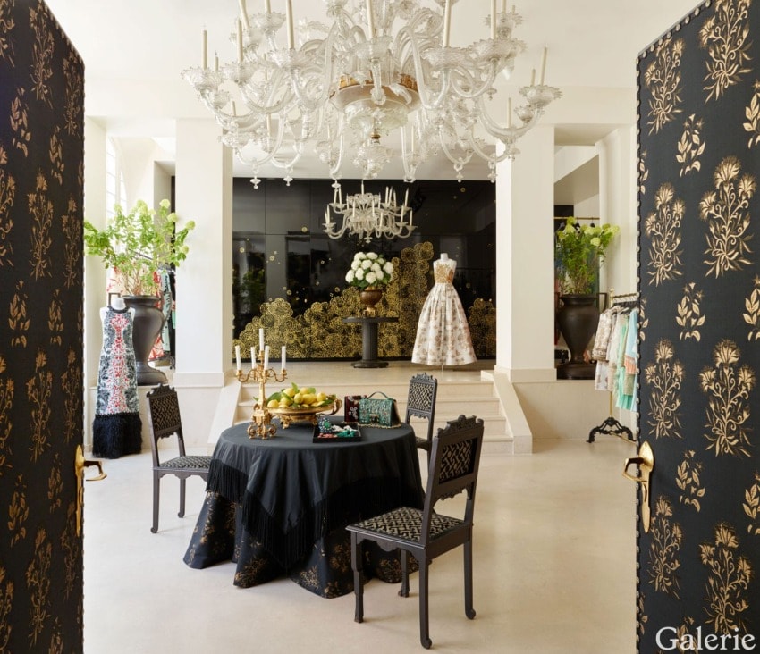Elegant dressing room with chandelier, luxurious gown display, decorative table with candles and fruit, surrounded by chairs.