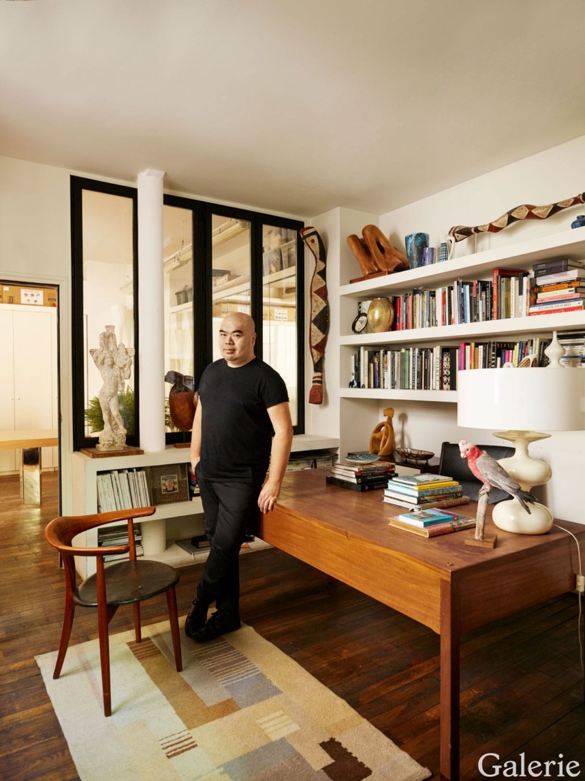 Person standing by a wooden desk in a stylish room with shelves filled with books and decorative items, wooden flooring.