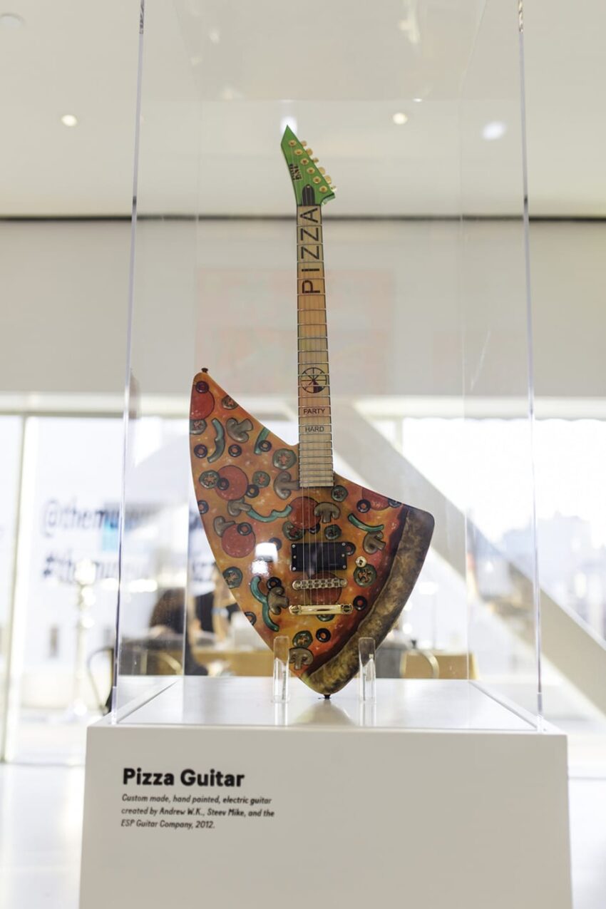 Unique guitar with pizza-themed design, showcased in glass display at a museum.