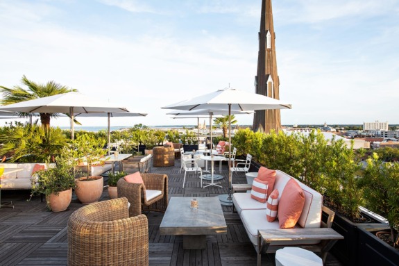 Rooftop patio with wicker furniture, tables, umbrellas, and lush greenery, overlooking a cityscape and distant horizon.