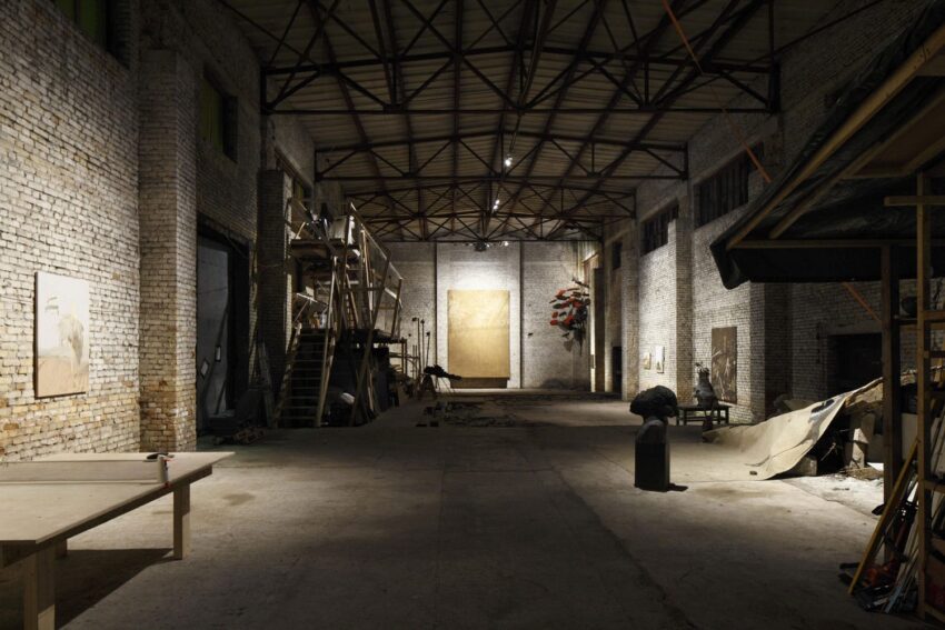 Industrial-style art gallery with exposed brick walls, high ceiling, and various art pieces displayed throughout the spacious interior.