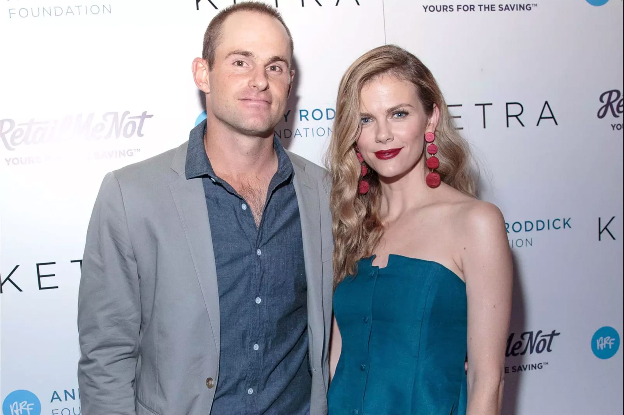 Man in gray suit and woman in blue dress with red earrings posing at an event with a step and repeat background.