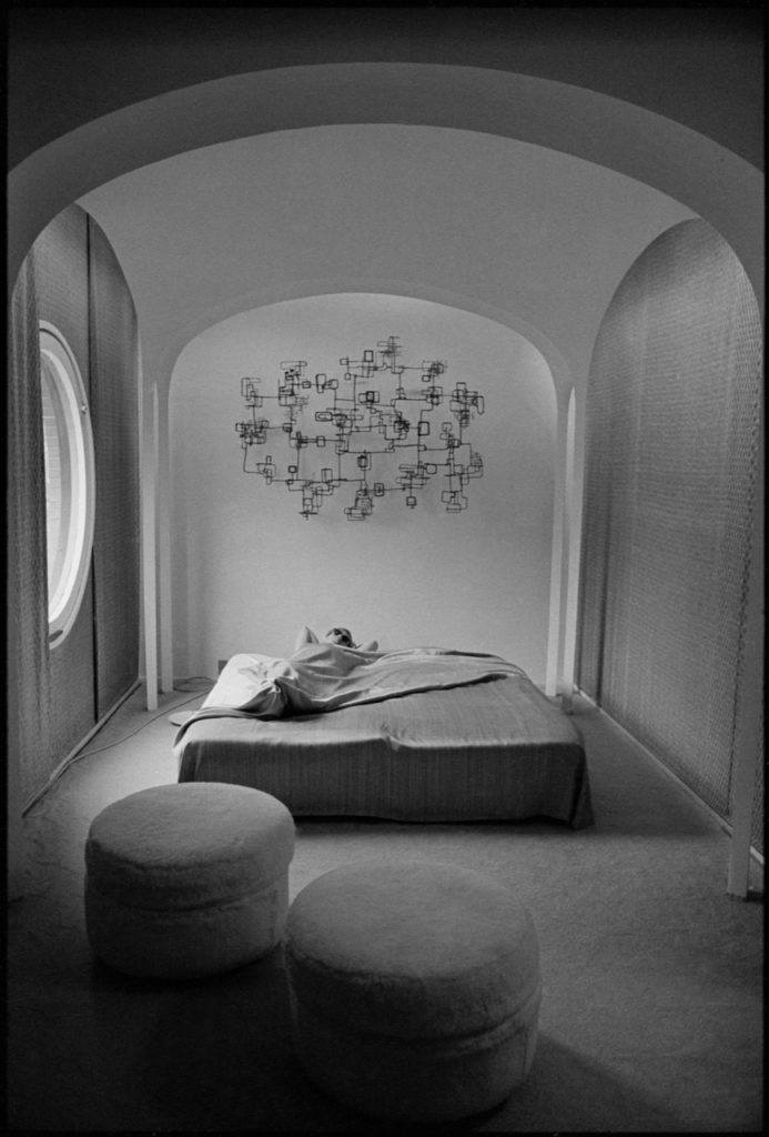 Minimalist bedroom with a circular window, abstract wall art, a low bed, and round ottomans on a carpeted floor.