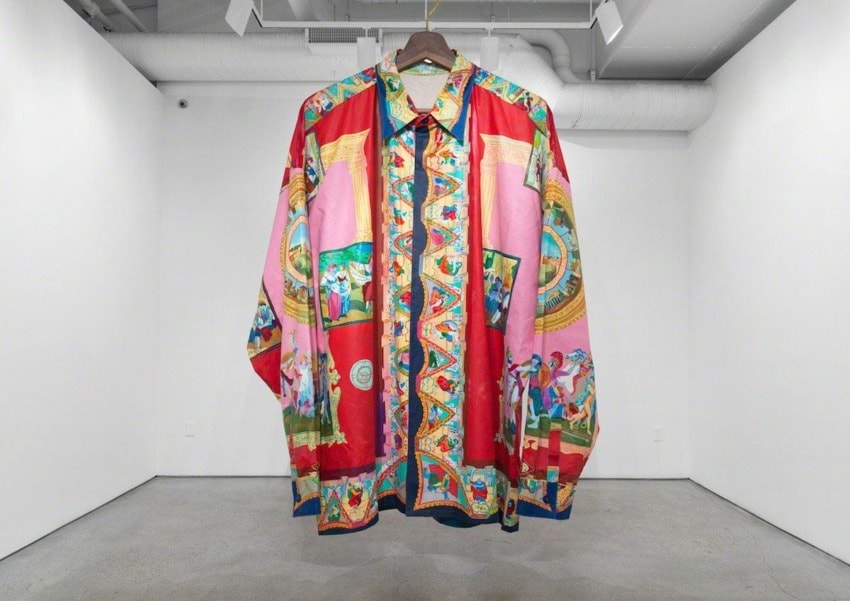 Colorful patterned shirt with intricate designs, hanging in a minimalist gallery space with white walls and a gray floor.