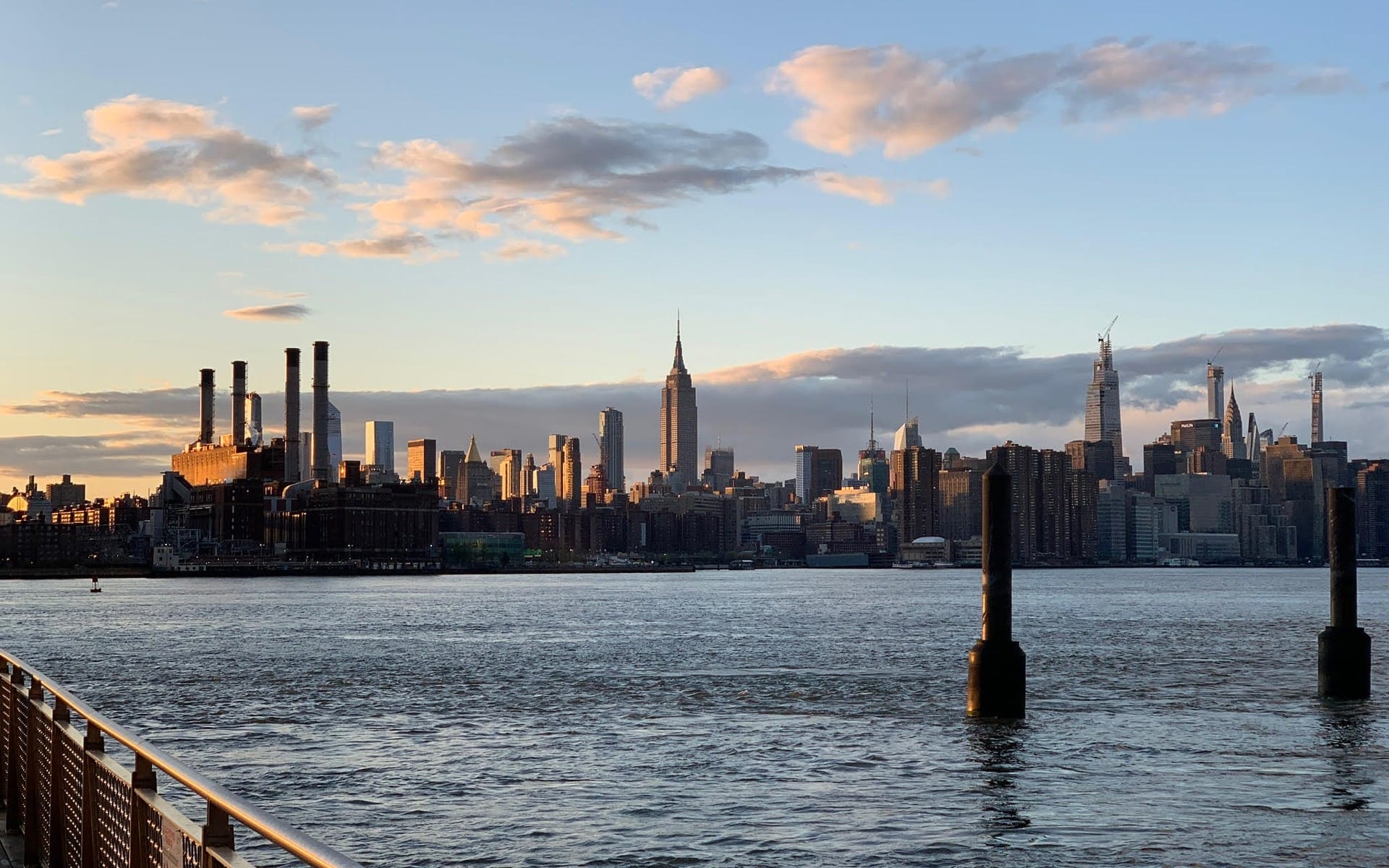 New York City skyline at sunset with water in the foreground and clouds in the sky.