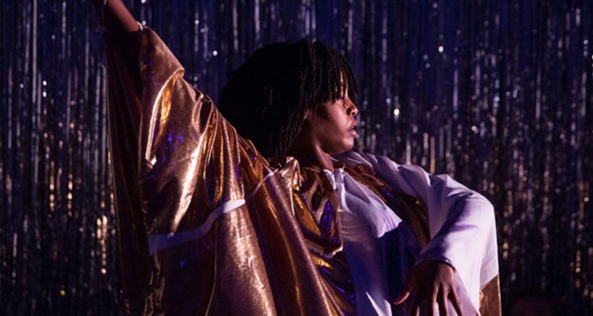 Person in gold and white costume performing dance with dramatic pose against a sparkling backdrop.