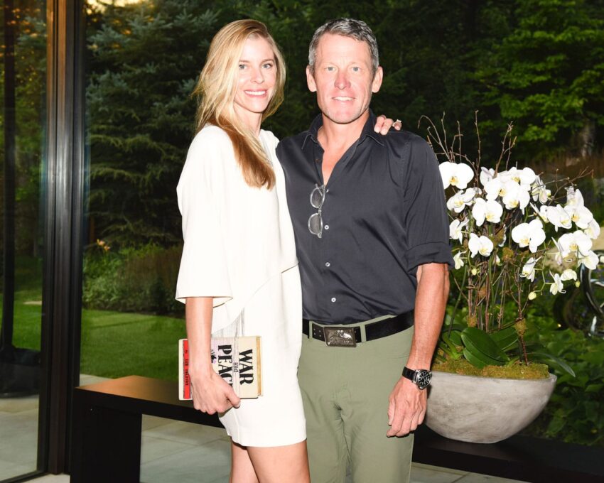 Smiling couple poses together indoors, woman holding book with orchids nearby, large windows showing greenery outside.