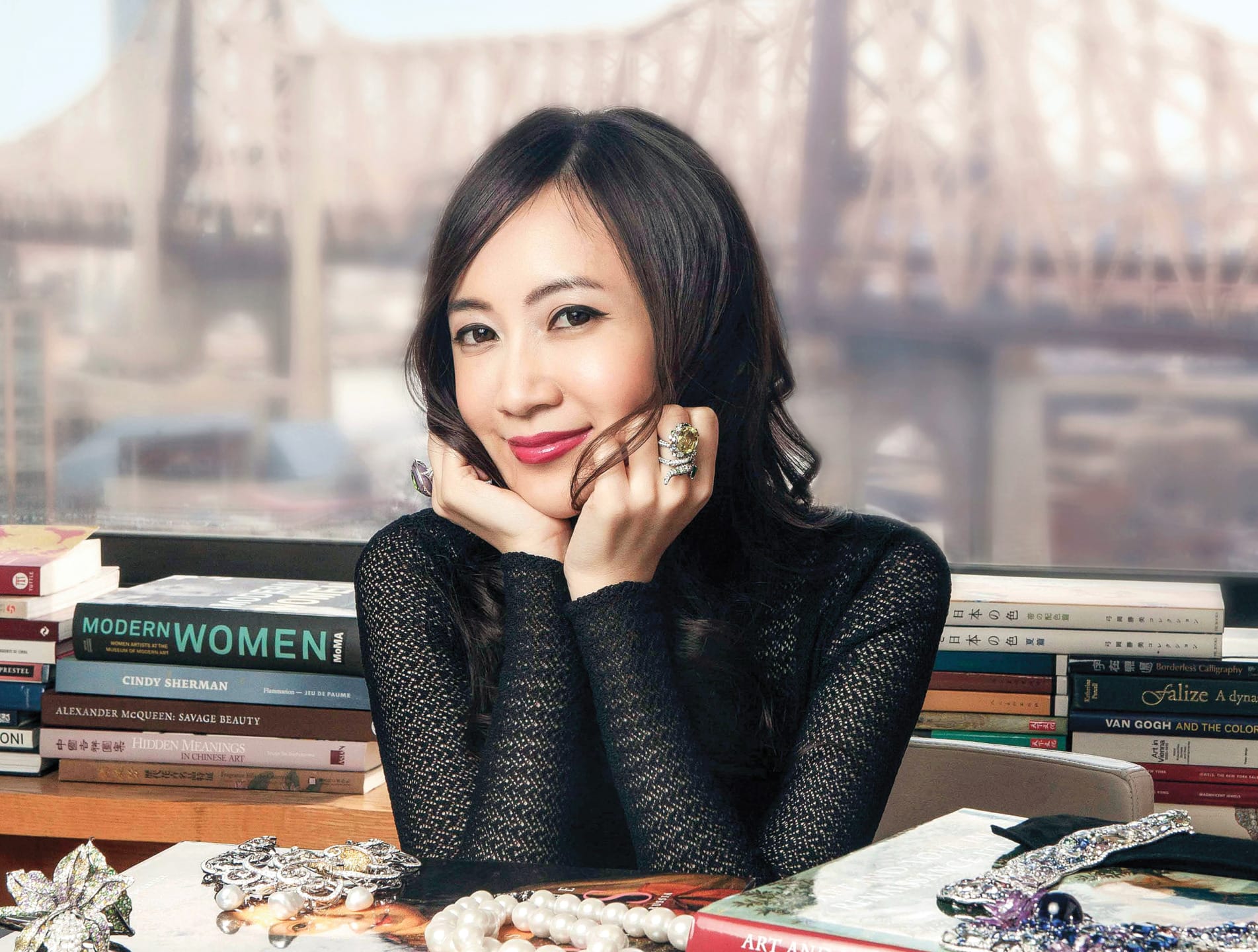 Smiling woman sitting at a desk with jewelry and books, a bridge visible in the background.