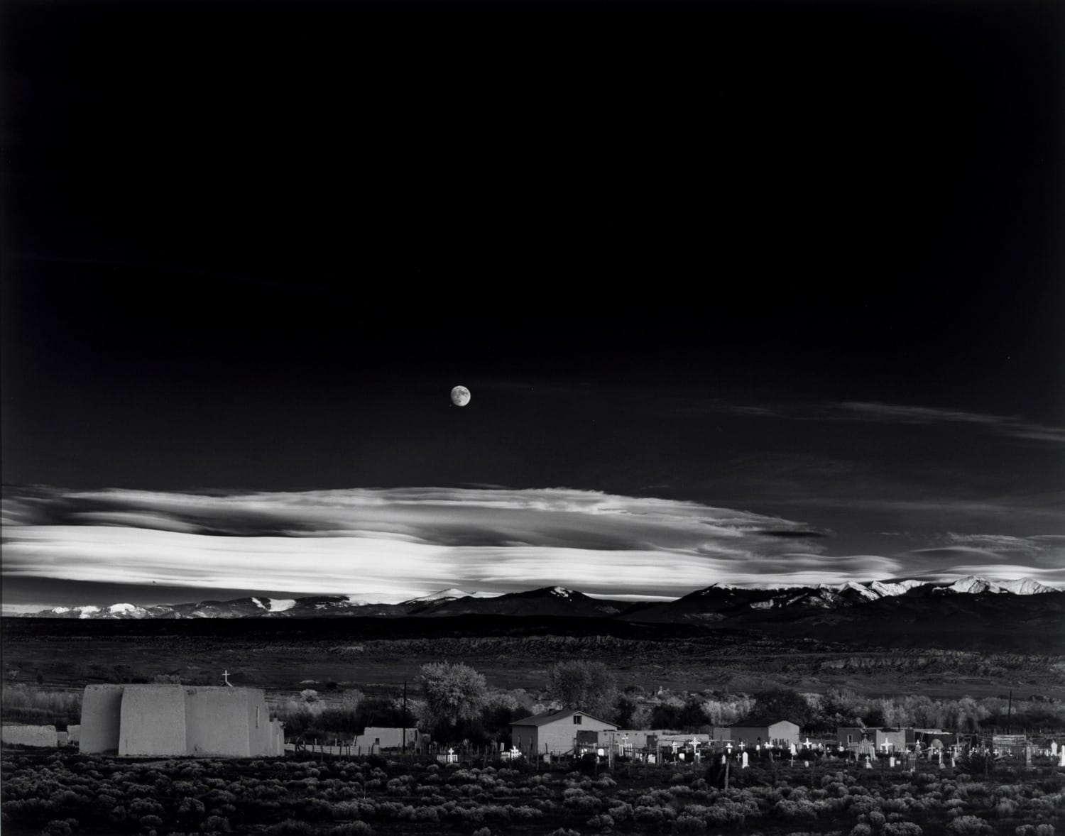 Black and white landscape with moon over distant mountains and scattered buildings under a dramatic cloudy sky.