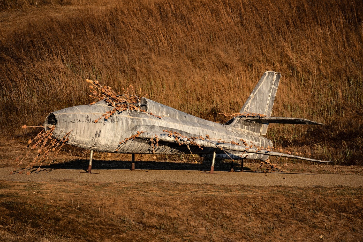 Rusty fighter jet sculpture covered in metallic spikes amidst dry grassy terrain.