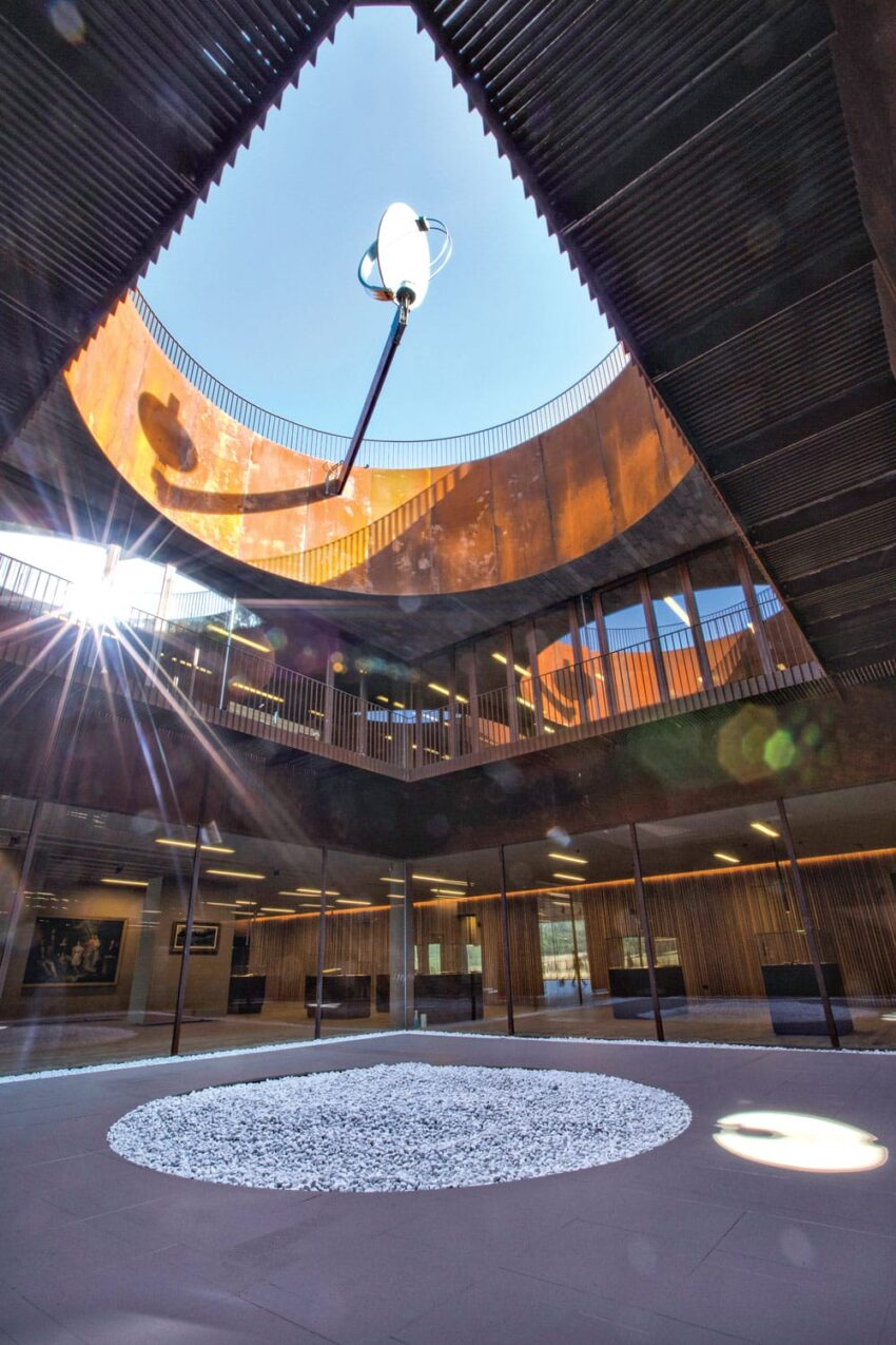 View of a modern atrium with circular skylight, wooden slatted walls, and sunlight creating lens flare on a white stone circle.