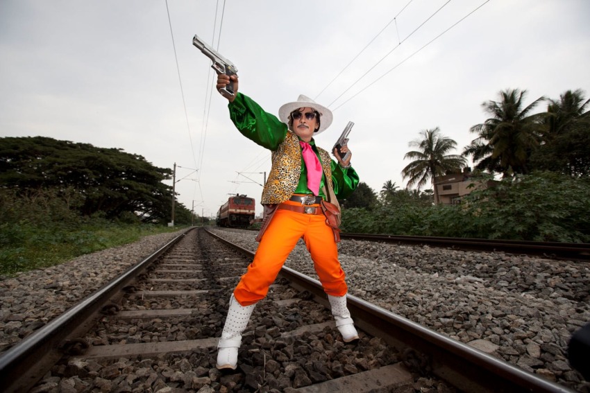 Person in colorful cowboy outfit poses with toy guns on railway tracks, trees and train in the background.