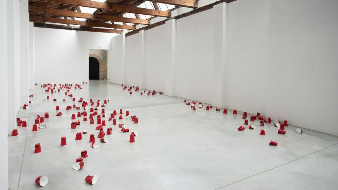Art installation with scattered red cups on a polished concrete floor in a bright gallery space with wooden beams and white walls.