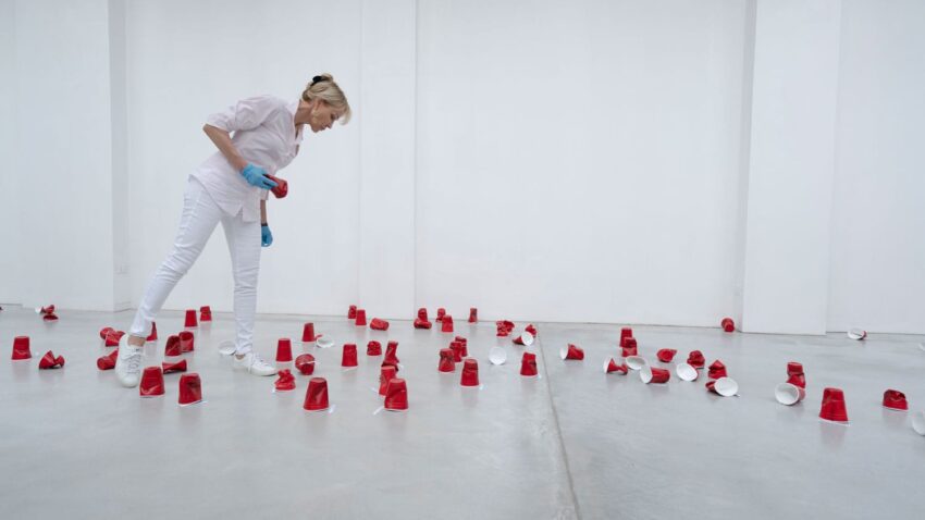 Person in white observing scattered red cups on a gallery floor.